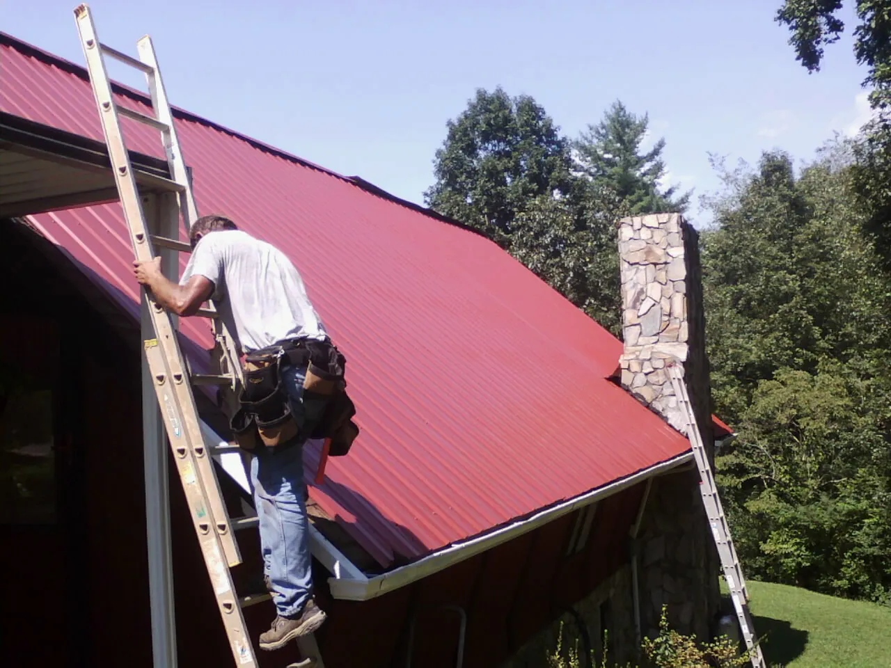 Roofer on a ladder beside a red metal roof and stone chimney — Soffit & Fascia Repair by Roofing Brothers in Leck Kill